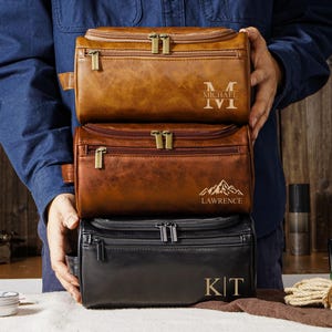 Puede incluir: Tres neceseres de cuero en tonos marrón y negro están apilados. La bolsa superior es de color marrón claro con "MICHAEL" e iniciales. La bolsa del medio es de color marrón más oscuro con "LAWRENCE" y un gráfico de montaña. La bolsa inferior es negra con las iniciales "KT".