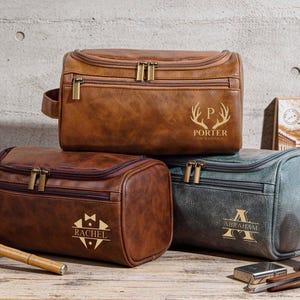 May include: Three brown and teal leather wash bags with gold zips and personalised text. The top bag is brown with the name "PORTER" and a deer antler design. The middle bag is brown with the name "RACHEL". The bottom bag is teal with the name "ABRAHAM".