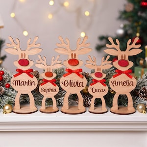 Könnte beinhalten: Fünf hellfarbene Holzrentier-Ornamente auf runden Sockeln. Jedes Rentier hat eine rote Nase, eine rote Schleife und einen auf den Körper gravierten Namen: Martin, Sophia, Olivia, Lucas und Amelia. Die Figuren haben unterschiedliche Größen.
