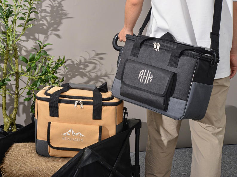 May include: Two insulated cooler bags. One is tan with black accents and a mountain logo with the name "Benjamin". The other is black and gray with a monogram. Both have top handles and shoulder straps.