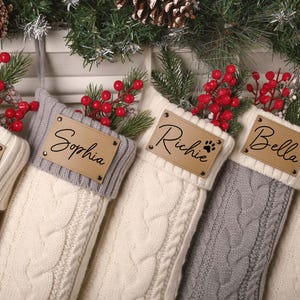 May include: Close-up of personalized Christmas stockings in various colors, including cream and gray, with cable knit design. Each stocking has a leather tag with a name and a paw print. Red berries and pine branches add festive detail.