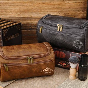 May include: Two leather wash bags, one brown and one grey, with gold zips. The brown bag has the name "BRENT" and a mountain graphic. The grey bag has the name "RICHARD". A shaving brush and bottle are also present.