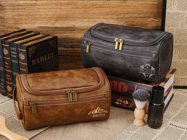 May include: Two leather wash bags, one brown and one grey, with gold zips. The brown bag has the name "BRENT" and a mountain graphic. The grey bag has the name "RICHARD". A shaving brush and bottle are also present.