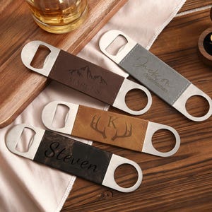 Peut inclure: Quatre ouvre-bouteilles personnalisés avec des têtes en acier inoxydable et des poignées en cuir. Les poignées sont marron, grises et noires, avec des noms, initiales et motifs de montagne et de bois gravés. Une boîte en bois avec des boutons de manchette dorés est en arrière-plan.