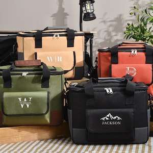 May include: A collection of insulated cooler bags in various colors: tan, orange, green, and black. Each bag features a front pocket with personalized text or initials. They have black handles, zippers, and shoulder straps, ideal for picnics or outdoor events.