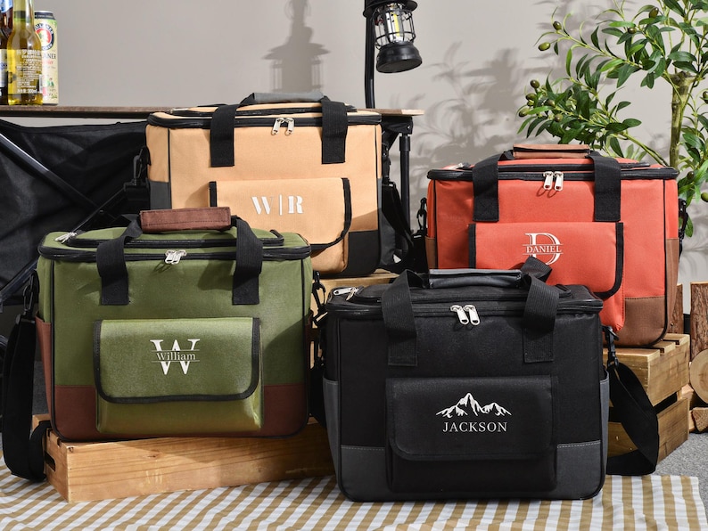 May include: A collection of insulated cooler bags in various colors: tan, orange, green, and black. Each bag features a front pocket with personalized text or initials. They have black handles, zippers, and shoulder straps, ideal for picnics or outdoor events.