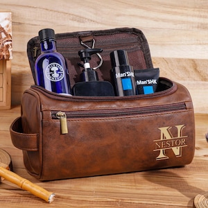May include: A brown leather wash bag with a gold zip and the name "Nestor" engraved on the front. Inside are grooming products, including a blue bottle and black bottles. A shaving brush and razor are also visible.