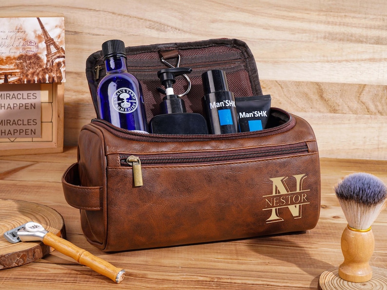 May include: A brown leather wash bag with a gold zip and the name "Nestor" engraved on the front. Inside are grooming products, including a blue bottle and black bottles. A shaving brush and razor are also visible.