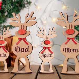 Könnte beinhalten: Weihnachtsdekorationen für eine Holz-Rentierfamilie. Jedes Rentier hat eine rote Nase und Schleife, mit Namen wie "Bella", "Dad", "Spike" und "Mom" auf ihren Körpern eingraviert. Die Rentiere stehen auf einer Holzoberfläche.