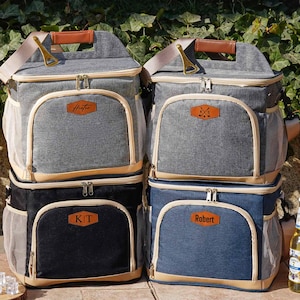 Könnte beinhalten: Vier personalisierte isolierte Kühltaschen mit braunen Lederelementen. Die Taschen sind paarweise gestapelt, wobei die beiden oberen Taschen grau und die beiden unteren Taschen schwarz und blau sind. Die Taschen haben Reißverschlüsse und Netzseitentaschen. Die Taschen sind mit den Namen "Hunter", "Kit", "Robert" und einem Symbol von zwei gekreuzten Golfschlägern beschriftet.