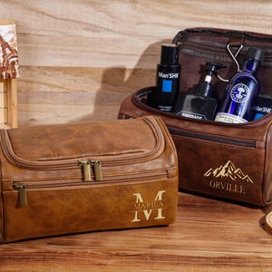 May include: Two brown leather wash bags with gold zips and handles. One bag is open, revealing bottles of grooming products. The other bag has the initials "M" and "I" with the Roman numeral "I". A wooden sign says "Miracles Happen".