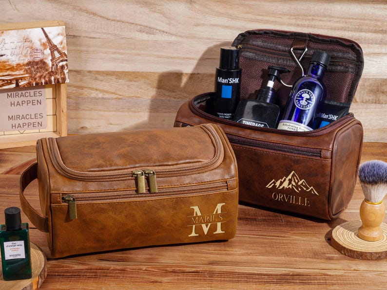 May include: Two brown leather wash bags with gold zips and handles. One bag is open, revealing bottles of grooming products. The other bag has the initials "M" and "I" with the Roman numeral "I". A wooden sign says "Miracles Happen".