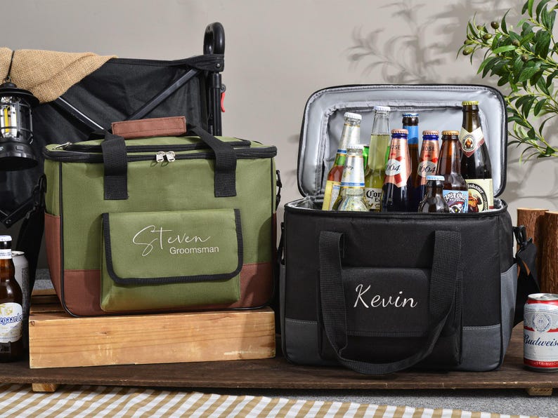 May include: Two insulated cooler bags, one olive green and one black, sit side-by-side. The green cooler has "Steven Groomsman" embroidered on the front. The black cooler has "Kevin" embroidered on the front. Both are open, revealing bottles of beer.