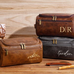 May include: Three personalized brown and black leather wash bags stacked on a wooden surface. The top bag has the initials "DR", the middle bag has the name "Gordon", and the bottom bag has the name "Francis" with a mountain graphic. A shaving brush and cut-throat razor are also visible.