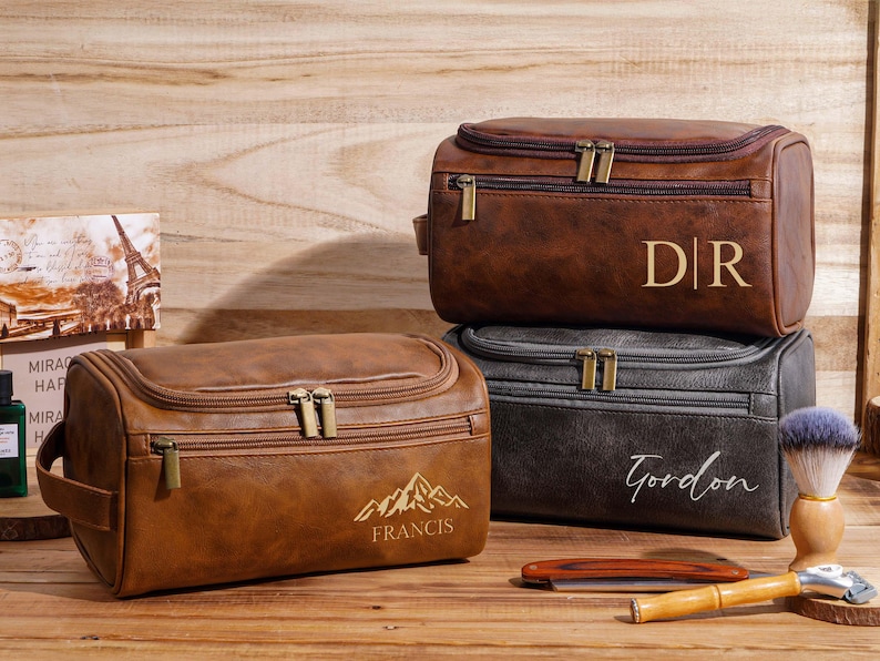 May include: Three personalized brown and black leather wash bags stacked on a wooden surface. The top bag has the initials "DR", the middle bag has the name "Gordon", and the bottom bag has the name "Francis" with a mountain graphic. A shaving brush and cut-throat razor are also visible.