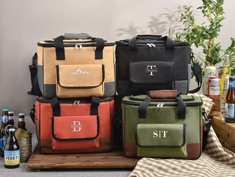 May include: Four personalized cooler bags in tan, black, red, and green. Each bag has a front pocket with a monogram or name. The bags have black handles, zippers, and a shoulder strap. The tan bag has a mountain design.