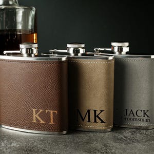 Puede incluir: Tres cantimploras de cuero personalizadas con iniciales grabadas. Las cantimploras son marrón, beige y gris. La cantimplora gris tiene el texto "JACK GROOMSMAN" grabado.