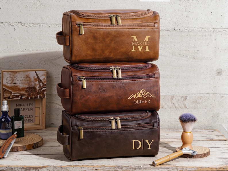 May include: Three brown leather wash bags stacked. Each has a zip closure and a side handle. The top bag has "MARTIN" and "IVI" in gold. The middle bag has a mountain graphic and "OLIVER". The bottom bag has "DIY". The bags are likely 25-30 cm long.