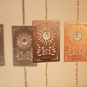 May include: Two gold-toned metal sun-shaped ornaments with the text "XIX. THE SUN" hanging from strings of small, light brown beads.