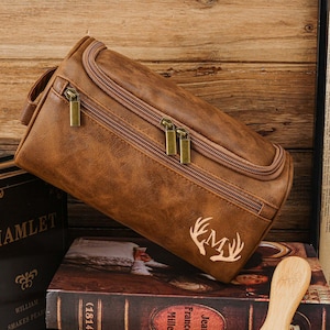 May include: A brown leather wash bag with a zip closure and a handle. The bag features a deer antler monogram and rests on a book. The bag is made of a textured material and has gold-coloured zips.