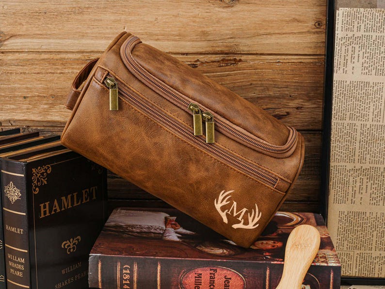 May include: A brown leather wash bag with a zip closure and a handle. The bag features a deer antler monogram and rests on a book. The bag is made of a textured material and has gold-coloured zips.