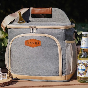 Peut inclure: Un sac isotherme gris avec une poignée en cuir marron et une fermeture à glissière. Le sac a un patch en cuir marron avec le nom "DAVID" dessus. Une bouteille de bière et des glaçons se trouvent devant le sac.