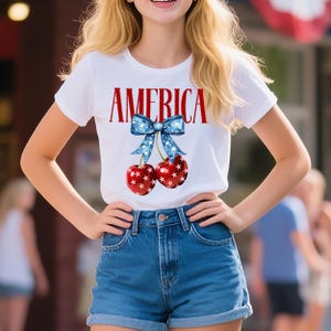May include: White t-shirt with the word "AMERICA" in red block letters. A blue bow with white stars is above two red cherries with white star patterns. The person is wearing blue denim shorts.