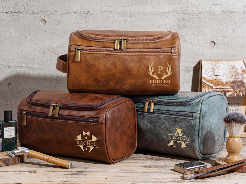 May include: Three brown and teal leather wash bags with gold zips and personalised text. The top bag is brown with the name "PORTER" and a deer antler design. The middle bag is brown with the name "RACHEL". The bottom bag is teal with the name "ABRAHAM".
