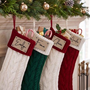 Könnte beinhalten: Vier Weihnachtsstrümpfe aus Zopfstrick in Rot, Grün und Weiß, jeweils mit einem rechteckigen Etikett in Lederoptik, auf dem die Namen Evelyn, Fiona, Claire und Amelia in schwarzer Schrift stehen. Jeder Strumpf enthält kleine Geschenke und Zuckerstangen.