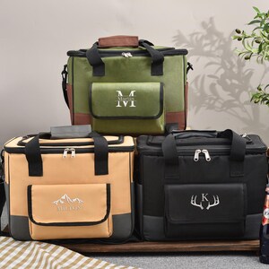 May include: Three insulated cooler bags in olive green, tan, and black, each with a front pocket and top handle. The olive green bag has a brown top and pocket trim, with the initials "M M" embroidered on the pocket. The tan bag has a mountain design and the name "Milton" embroidered on the pocket. The black bag has a deer antler design and the letter "K" embroidered on the pocket.