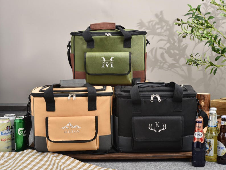 May include: Three insulated cooler bags in olive green, tan, and black, each with a front pocket and top handle. The olive green bag has a brown top and pocket trim, with the initials "M M" embroidered on the pocket. The tan bag has a mountain design and the name "Milton" embroidered on the pocket. The black bag has a deer antler design and the letter "K" embroidered on the pocket.
