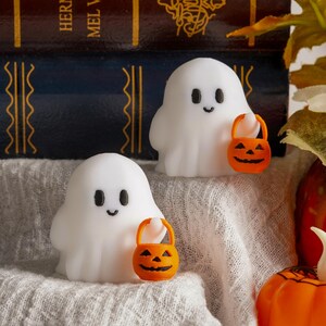 Könnte beinhalten: Zwei weiße Geisterfiguren mit schwarzen Augen und Lächeln, die jeweils einen orangefarbenen Kürbis-Eimer halten. Die Geister stehen auf einem weißen Tuch, mit Büchern und Herbstdekorationen im Hintergrund. Halloween-Dekoration.