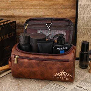 May include: A brown leather wash bag with a zip, containing grooming products. The bag has a mountain and "MARTIN" design. Other items include a shaving brush, a bottle, and books.