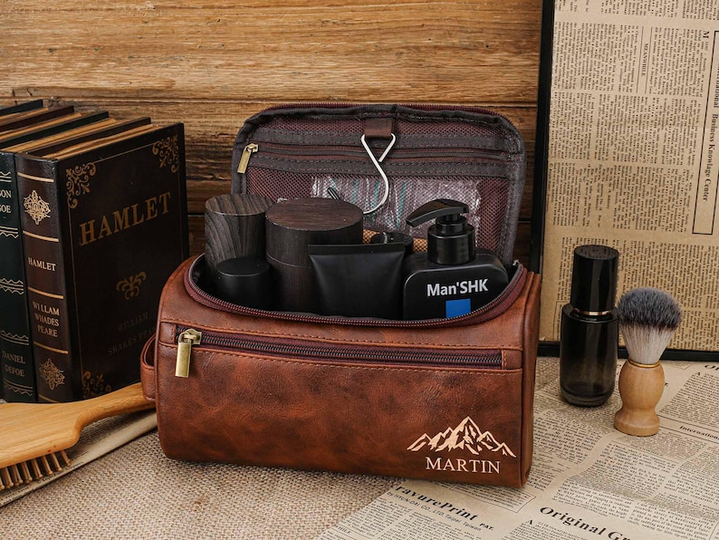 May include: A brown leather wash bag with a zip, containing grooming products. The bag has a mountain and "MARTIN" design. Other items include a shaving brush, a bottle, and books.