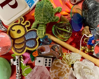 May include: A collection of small decorative items on a red surface. Includes a green turtle figurine, a butterfly patch, a seashell, a die, a key charm, a pink crystal, and a stamp from Italy. Also present are feathers, beads, and a wooden stick.