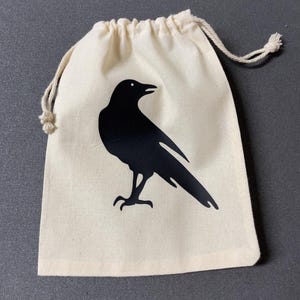 May include: A natural canvas drawstring bag featuring a black silhouette of a crow. The bag has a beige drawstring closure and is made of a soft, durable fabric. The crow design is centered on the front of the bag.