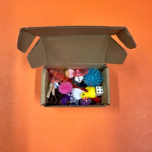 May include: A small, open cardboard box filled with an assortment of colorful trinkets. Items include a yellow rubber duck, dice, a blue spiky ball, pink pom-poms, and other small decorative objects. The box sits on an orange background.