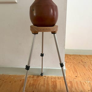 May include: A brown ceramic vase sits atop a wooden stand with the word "ORBIS" printed on it. The stand is supported by a silver tripod with black adjustment clips. The vase has a rounded body and a narrow neck.