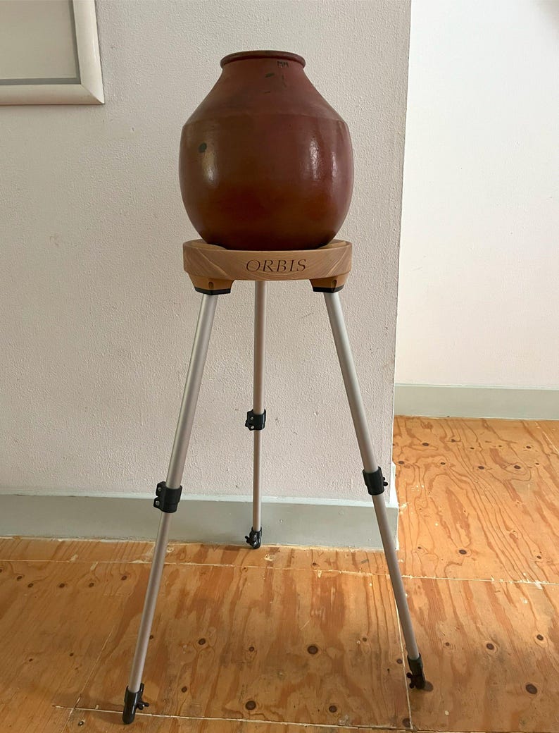 May include: A brown ceramic vase sits atop a wooden stand with the word "ORBIS" printed on it. The stand is supported by a silver tripod with black adjustment clips. The vase has a rounded body and a narrow neck.