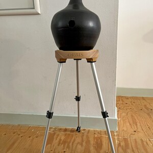 May include: A black ceramic vase with a narrow neck and a round body, supported by a wooden stand with the word "ORBIS" printed on it. The stand is mounted on a silver tripod with adjustable legs.