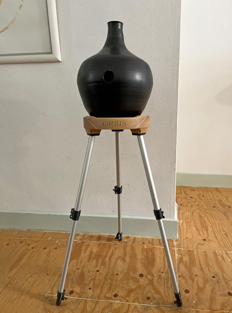 May include: A black ceramic vase with a narrow neck and a round body, supported by a wooden stand with the word "ORBIS" printed on it. The stand is mounted on a silver tripod with adjustable legs.
