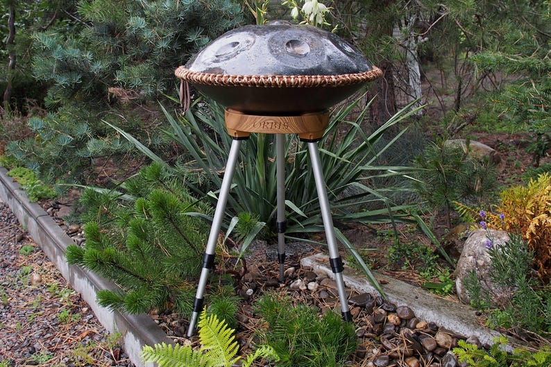 May include: A dark gray steel handpan drum with a brown woven trim, mounted on a wooden stand with three silver legs. The instrument is set outdoors amidst lush greenery.