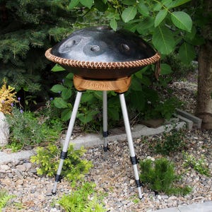 May include: A black steel handpan drum with a brown leather trim, resting on a wooden stand with the word "VIRUS" printed on it. The stand is supported by a silver tripod. The instrument is set outdoors among greenery.
