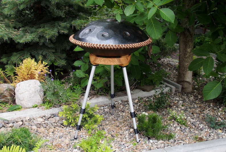 May include: A black steel handpan drum with a brown leather trim, resting on a wooden stand with the word "VIRUS" printed on it. The stand is supported by a silver tripod. The instrument is set outdoors among greenery.