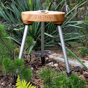 Könnte beinhalten: Ein Holzstativ mit silbernen Beinen und einer kreisförmigen Oberseite. Das Wort "ORBIS" ist auf die Holzoberfläche graviert. Das Stativ steht in einem Garten mit grünen Pflanzen und Steinen.