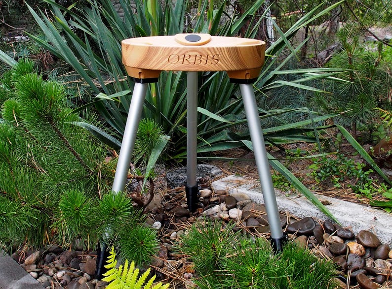 May include: A wooden tripod with silver legs and a circular top. The word "ORBIS" is engraved on the wooden surface. The tripod is set in a garden with green plants and rocks.