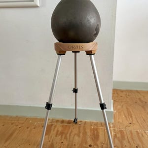 May include: A dark gray, egg-shaped ceramic vase sits atop a wooden stand with the word "ORBIS" engraved on it. The stand has three silver legs with black adjustable joints. The vase has a textured surface and a dark rim.