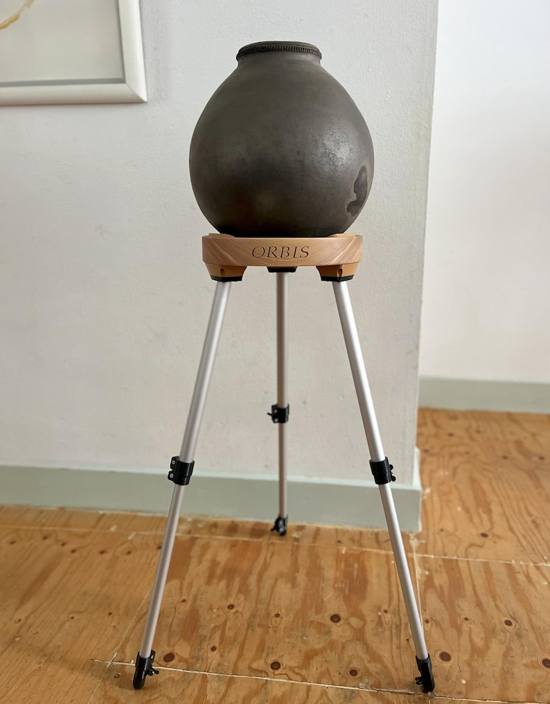 May include: A dark gray, egg-shaped ceramic vase sits atop a wooden stand with the word "ORBIS" engraved on it. The stand has three silver legs with black adjustable joints. The vase has a textured surface and a dark rim.