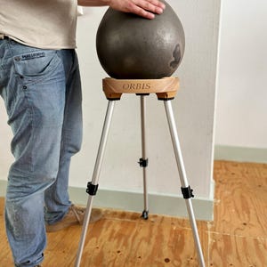 May include: A dark gray, teardrop-shaped sculpture rests on a wooden stand with the word "ORBIS" printed on it. The stand has three silver legs. The sculpture is being touched by a person's hand.