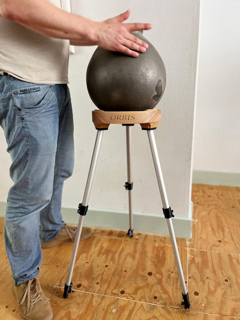 May include: A dark gray, teardrop-shaped sculpture rests on a wooden stand with the word "ORBIS" printed on it. The stand has three silver legs. The sculpture is being touched by a person's hand.